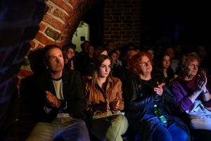 Koncert: Pracownia piosenki prezentuje - Niepozorny zaścianek - Zdjęcie pokazuje widownię w Piwnicach TCK podczas koncertu. Widzowie, siedzący na krzesłach obserwują występ. Na zdjęciu widać różnorodność publiczności, od młodych po starszych słuchaczy. Sala jest delikatnie oświetlona.