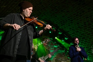 Koncert jazzowy: Tomasz Chyła Quintet - Zdjęcie sceny z koncertu Tomasz Chyła Quintet, pokazujące zaangażowanych muzyków. Tomasz Chyła gra na skrzypcach, w tle pozostali muzycy na różnych instrumentach. Scena jest dynamicznie oświetlona, co dodaje energii występowi.