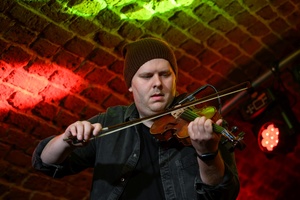 Koncert jazzowy: Tomasz Chyła Quintet - Zdjęcie sceny z koncertu Tomasz Chyła Quintet, pokazujące zaangażowanych muzyków. Tomasz Chyła gra na skrzypcach, w tle pozostali muzycy na różnych instrumentach. Scena jest dynamicznie oświetlona, co dodaje energii występowi.