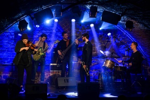 Koncert jazzowy: Tomasz Chyła Quintet - Zdjęcie sceny z koncertu Tomasz Chyła Quintet, pokazujące zaangażowanych muzyków. Tomasz Chyła gra na skrzypcach, w tle pozostali muzycy na różnych instrumentach. Scena jest dynamicznie oświetlona, co dodaje energii występowi.