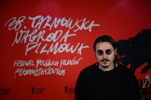 38. TNF - Dzień 2 (1.06.2024) - Maks Bereski, autor plakatu artystycznego 38. TNF na tle ścianki festiwalowej w rozmowie do kroniki festiwalowej